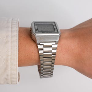 May include: A silver digital watch with a rectangular face and a matching metal band. The watch displays the time and date. The watch is on a wrist, and the background is a neutral color.