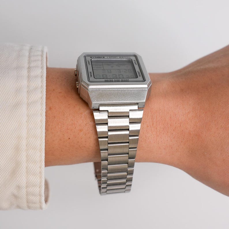 May include: A silver digital watch with a rectangular face and a matching metal band. The watch displays the time and date. The watch is on a wrist, and the background is a neutral color.