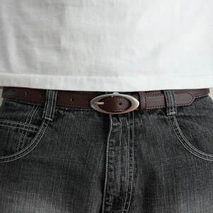 May include: A brown leather belt with a silver oval buckle is worn with dark denim jeans. The belt is secured around the waist, with the buckle centered. The jeans have white stitching.