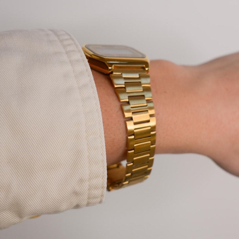May include: A gold-toned wristwatch with a rectangular face and a matching link band. The watch is worn on a wrist, with a portion of a light-colored long-sleeved shirt visible in the frame. The watch band is approximately 7 inches in length.