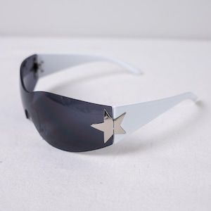 May include: White sunglasses with dark gray lenses. The sunglasses feature a silver star accent on the outer corners. The arms are white and curve slightly. The sunglasses are set against a white background.