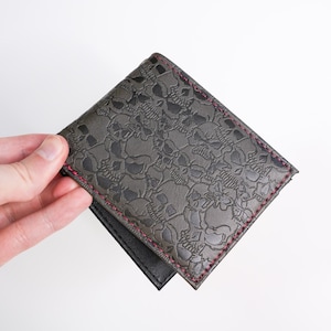 May include: A bi-fold wallet with a dark gray skull pattern embossed on the front. The wallet has a black interior and is stitched with pink thread around the edges. The wallet is held open, revealing the interior.