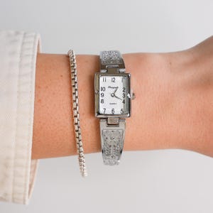 May include: A silver-toned rectangular watch with a white face and black numerals. The watch band and face frame feature an ornate, engraved design. A silver bracelet is visible next to the watch.