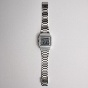 May include: A silver digital watch with a stainless steel band. The watch face displays the time, date, and a world map icon. The watch band is made of small, rectangular links. The watch is displayed on a white background.
