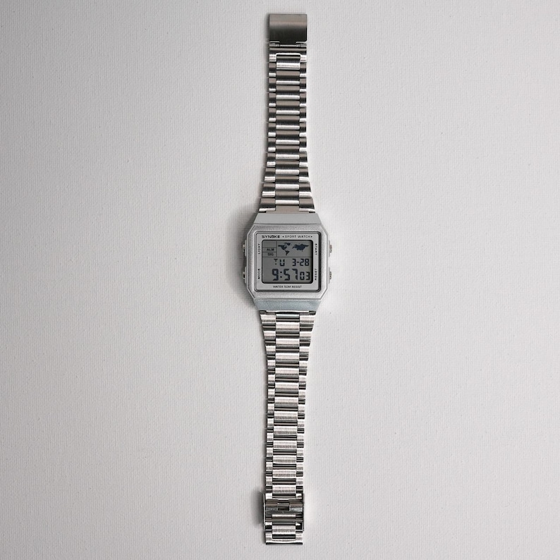 May include: A silver digital watch with a stainless steel band. The watch face displays the time, date, and a world map icon. The watch band is made of small, rectangular links. The watch is displayed on a white background.