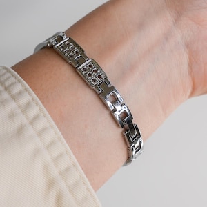 May include: A silver-toned bracelet with a geometric design. The bracelet features rectangular links with small, red-colored accents. The bracelet is worn on a wrist, showcasing its design and color.