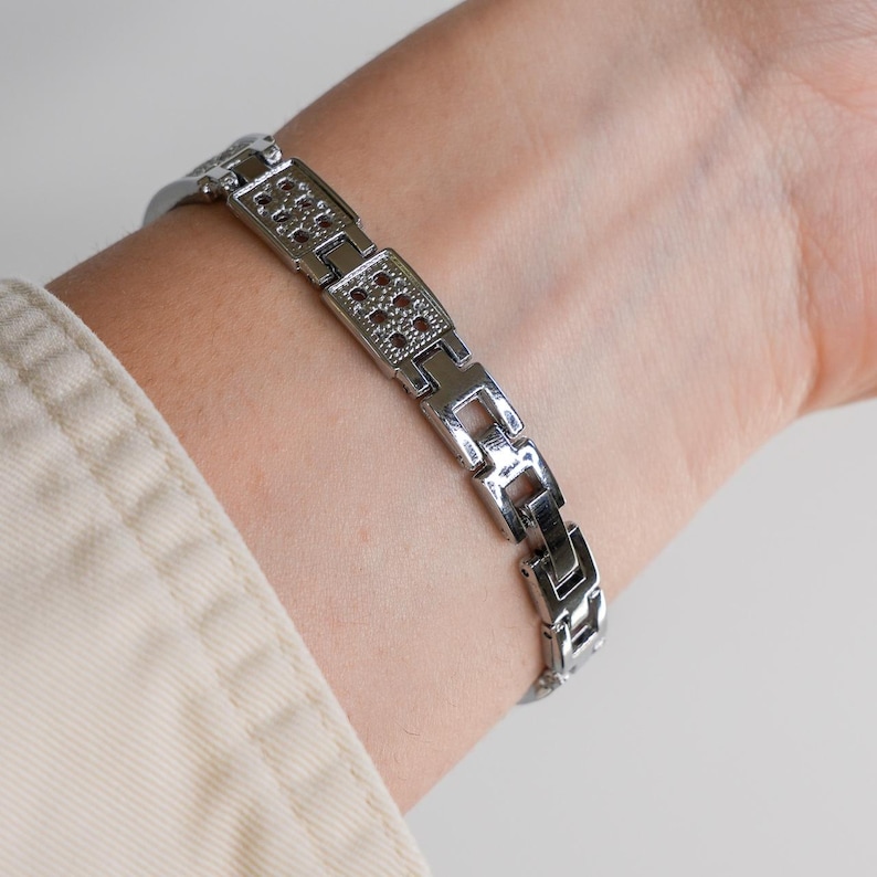 May include: A silver-toned bracelet with a geometric design. The bracelet features rectangular links with small, red-colored accents. The bracelet is worn on a wrist, showcasing its design and color.