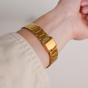 May include: A gold-colored watch band is fastened around a wrist. The watch band is made of rectangular links and has a rectangular clasp. The wrist is partially visible, and the person is wearing a light-colored shirt.