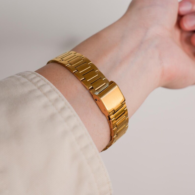 May include: A gold-colored watch band is fastened around a wrist. The watch band is made of rectangular links and has a rectangular clasp. The wrist is partially visible, and the person is wearing a light-colored shirt.