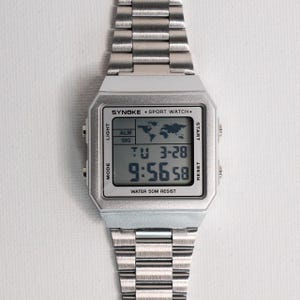 May include: A silver digital sport watch with a rectangular face and a stainless steel band. The watch face displays the time, date, and world map icon. The text "SYNOKE SPORT WATCH" and "WATER 50M RESIST" are visible.