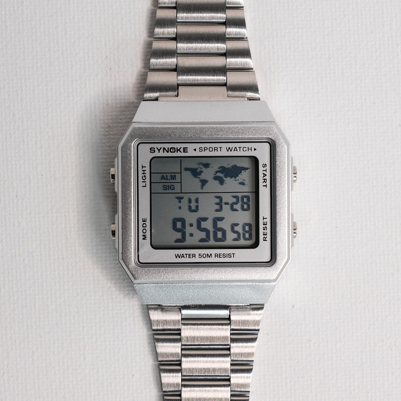 May include: A silver digital sport watch with a rectangular face and a stainless steel band. The watch face displays the time, date, and world map icon. The text "SYNOKE SPORT WATCH" and "WATER 50M RESIST" are visible.