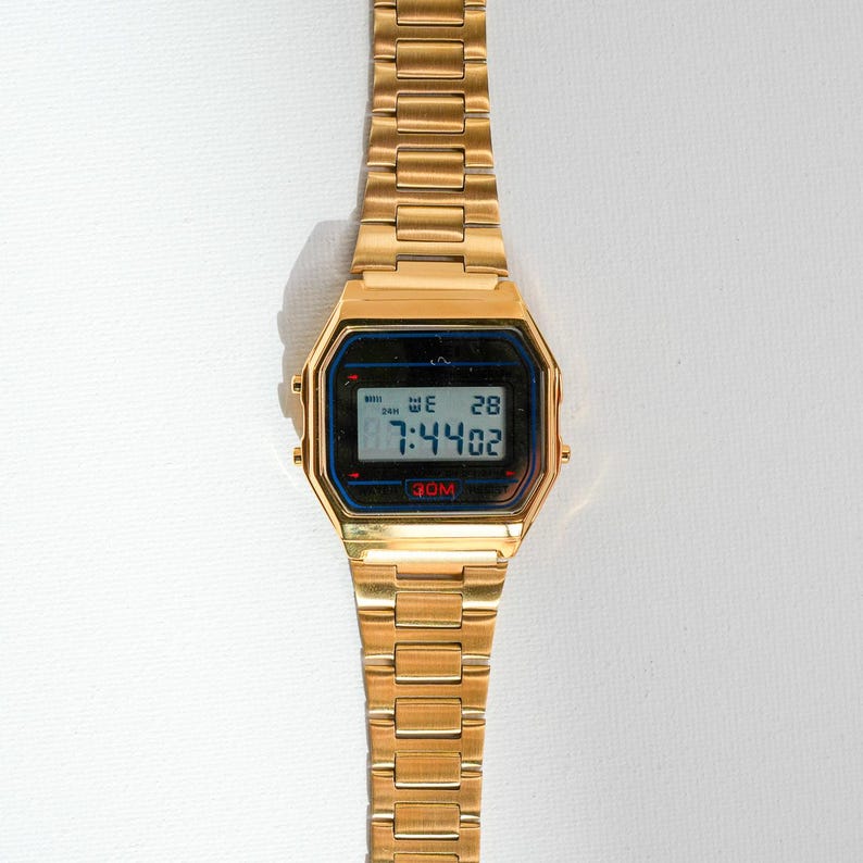 May include: A gold-toned digital watch with a rectangular face and a matching link band. The watch face displays the time, day, and date in a digital format. The watch is set against a white background.