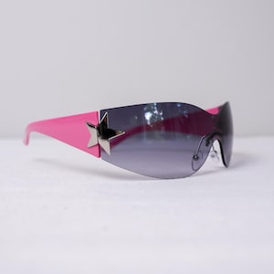 May include: Pink sunglasses with a dark gray gradient lens. The sunglasses feature a silver star accent on the side and pink arms. The overall design is sleek and modern.