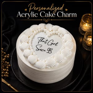 May include: A round white cake topped with a heart-shaped acrylic cake charm featuring the black text That Girl Since 93. The cake is decorated with white piped frosting shells around the perimeter of the heart-shaped center piece.