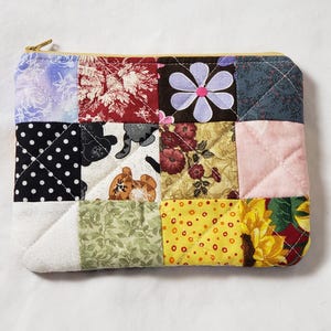 May include: A patchwork zippered pouch with a variety of fabric squares in floral, polka dot, and patterned designs. The pouch features a gold-colored zipper and is quilted for added texture and durability. The color palette includes pink, yellow, and blue.