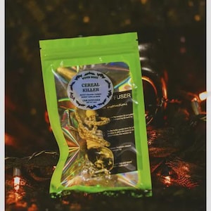May include: A green, transparent package with a car air freshener inside. The package has a label that reads "Cereal Killer" with a skull and bat design. The background is dark with string lights.