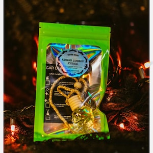 May include: A green, holographic-effect package containing a car air freshener. The package features a sticker with the text "Sugar Cookie Curse" and "Car Diffuser". The air freshener has a wooden bead and a decorative element.