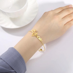 Op de afbeelding: Een goudkleurige, gepersonaliseerde naamarmband. De armband heeft de naam "Yecca" in een cursief lettertype, met een kleine kroon boven de eerste letter. De armband heeft een delicaat, open manchetontwerp.