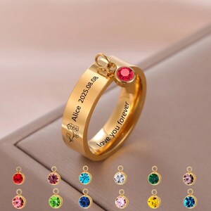 Könnte beinhalten: Ein goldfarbener Ring mit einem roten Edelsteinanhänger. Der Ring ist mit dem Namen "Alice", dem Datum "2025.08.08" und dem Satz "love you forever" graviert. Zusätzliche Edelsteinanhänger in verschiedenen Farben werden darunter angezeigt.