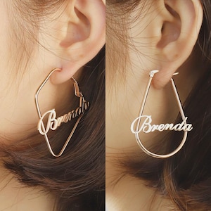 Op de afbeelding: Twee roségouden oorringen, één vierkant en één druppelvormig, elk met de naam "Brenda" in cursief schrift. De oorbellen worden op een oor getoond.