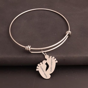 May include: A silver-colored bangle bracelet with a charm featuring two baby footprints. The charm has the words "custom name" engraved on it. The bracelet is made of a shiny metal and is displayed on a dark surface.