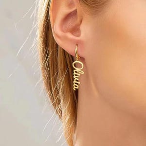Peut inclure: Une boucle d'oreille dorée avec le nom "Olivia" en écriture cursive. La boucle d'oreille est suspendue à l'oreille d'une personne. Le nom est aligné verticalement et suspendu à la boucle.