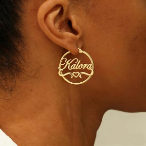 Puede incluir: Pendiente de aro dorado con el nombre "Kalora" en fuente cursiva. El nombre está centrado dentro del aro, con dos pequeños corazones debajo. El pendiente se lleva en la oreja.