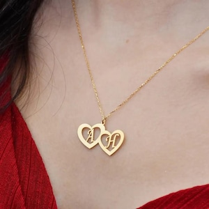 May include: A gold-colored necklace featuring two heart-shaped pendants. Each heart has a letter cut out, with the letters "A" and "H" visible. The necklace is worn against a red top.