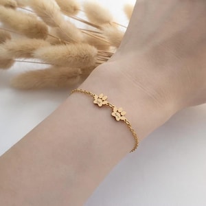 Könnte beinhalten: Goldfarbenes Armband mit zwei Pfotenabdruck-Anhängern. Jeder Anhänger hat einen eingravierten Namen. Das Armband wird am Handgelenk getragen, mit getrockneten Pflanzen im Hintergrund. Das Armband ist ein zartes Schmuckstück.