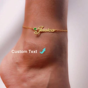 Può includere: Una cavigliera color oro con il nome "Jessica" in corsivo. La cavigliera presenta una piccola gemma verde ed è attaccata a una delicata catena. Il testo "Custom Text" è anche visibile.