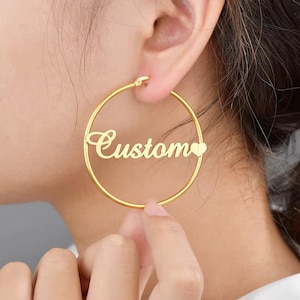 May include: Gold-colored hoop earrings with the word "Custom" in a cursive font. A small heart shape is on the right side of the word. The earrings are round and are being held up to an ear.