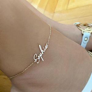 May include: A delicate gold anklet featuring the name "Sophia" in cursive. The anklet is worn on an ankle, with a white sandal visible. The fine chain adds to the elegant design of the personalised jewellery.