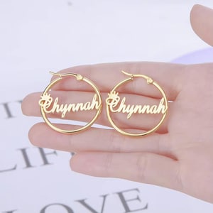 Könnte beinhalten: Ein Paar goldfarbene Creolen. Jeder Ohrring trägt den Namen "Chynnah" in kursiver Schrift, mit einem kleinen Kronen-Design. Die Ohrringe werden in einer Hand vor einem weichen Hintergrund gehalten.