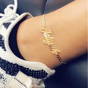 Può includere: Una cavigliera color oro con il nome "Jaliyah" in corsivo. La cavigliera presenta un design a catena a maglia. L'immagine mostra la cavigliera indossata su una caviglia, con una scarpa a fantasia bianca e nera sullo sfondo.