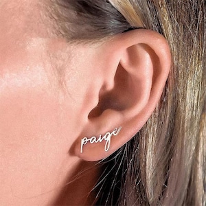 May include: A close-up of an ear with a silver earring spelling the name "Paige" in a cursive font. The earring is a delicate, minimalist design, perfect for everyday wear. The skin tone is light.