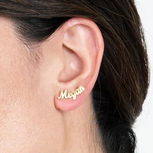 May include: Gold-coloured stud earrings spelling out the name "Megan" in a cursive script. The earrings are displayed on an ear, showcasing the personalised jewellery's design and fit. The earrings are a stylish accessory.