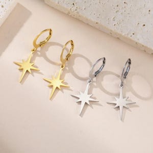 May include: Four earrings with starburst pendants. Two are gold-colored, and two are silver-colored. Each earring has a hoop closure. The earrings are displayed on a light pink surface.