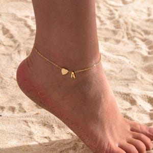 May include: A gold anklet featuring a heart pendant and the letter "A". The chain is worn on an ankle, with a sandy beach in the background. The anklet is a simple, elegant piece of jewellery.