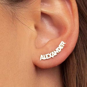 Może przedstawiać: Srebrny kolczyk typu ear climber z napisem "Alexander" w stylizowanej czcionce. Kolczyk dopasowany do kształtu ucha, z nazwą podążającą za krzywizną.