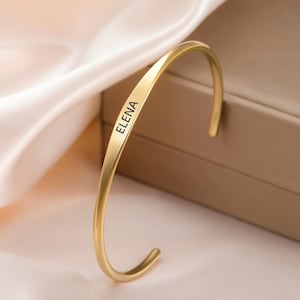 Puede incluir: Una pulsera de brazalete dorada y curvada. El nombre "ELENA" está grabado en negro en la parte delantera de la pulsera. La pulsera tiene un acabado liso y pulido y está sobre un fondo neutro.