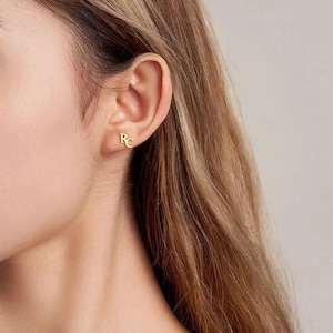 Puede incluir: Primer plano de una oreja con un pendiente de oro. El pendiente presenta las iniciales "RC" en una fuente cursiva delicada. El pendiente es pequeño y se encuentra cerca del lóbulo de la oreja.