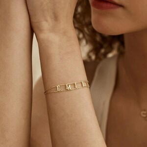 Puede incluir: Pulsera dorada con el nombre "EMILY" en letras mayúsculas. La pulsera es delicada, con un diseño de cadena. Las letras están conectadas y se colocan en la muñeca. La pulsera se lleva en el brazo de una persona.