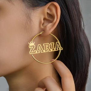 Könnte beinhalten: Goldfarbene Creolen mit dem Namen "ZARIA" in einer kräftigen Schrift, verziert mit einer kleinen Krone. Die kreisförmigen Ohrringe werden getragen.