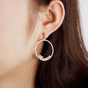 Könnte beinhalten: Roségoldene Creolen mit dem Namen "Blanca" in Schreibschrift. Die Ohrringe sind kreisförmig mit einem kleinen Stecker oben zur Befestigung. Das Namensdesign ist in den Reifen integriert.