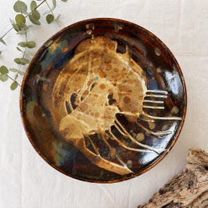 May include: A handcrafted ceramic plate with a distinctive design. The plate has a brown and tan colour scheme with blue and white accents. The design features an abstract, organic shape. The plate is round and appears glazed.