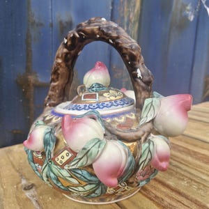 Könnte beinhalten: Dekorative Keramik-Teekanne mit braunem Griff und pfirsichförmigen Verzierungen. Die Teekanne hat einen goldenen Sockel mit bunten Blumen- und Blattmotiven sowie rosa und weißen Pfirsichdekorationen. Der Deckel ist mit einem pfirsichförmigen Knauf versehen.