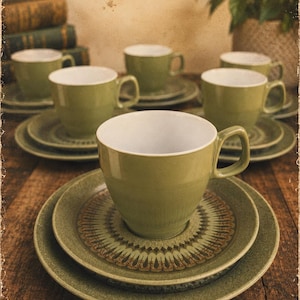 May include: A set of sage green ceramic cups and saucers featuring a white interior and a decorative brown and green radial pattern on the saucers. Each cup has a single handle and sits on a matching saucer, with multiple sets arranged in a group.