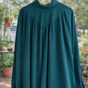 May include: A teal long-sleeve blouse with a high neckline and gathered detailing. The blouse is hanging on a wooden hanger. The sleeves have buttoned cuffs. The background is blurred, with greenery visible.