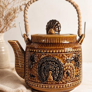 May include: A brown glazed ceramic teapot featuring a relief design of a peacock with a fanned tail. The teapot has a textured surface, a lid with a shell-shaped finial, and a curved handle wrapped in light-coloured woven material attached to metal loops.