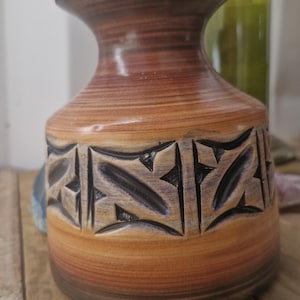 Può includere: Un vaso in ceramica marrone e beige con bordo blu scuro e design geometrico intagliato al centro. Il vaso ha una base bulbosa e un collo stretto, con una bottiglia sullo sfondo.